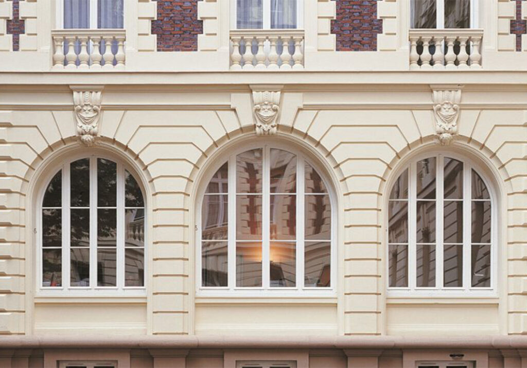 Zoom sur trois fenêtres cintrées de style haussmannien avec croisillons blancs, ornées de sculptures murales et de moulures, sur une façade en grès beige. 