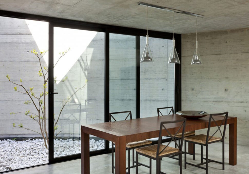 Salle à manger de type caveau, à l’ambiance feutrée, avec une décoration minimaliste dans les tons noirs et bois. La pièce est installée devant un mur rideau de plain-pied, composé de grandes menuiseries rectangulaires noires laissant entrer la lumière naturelle.
