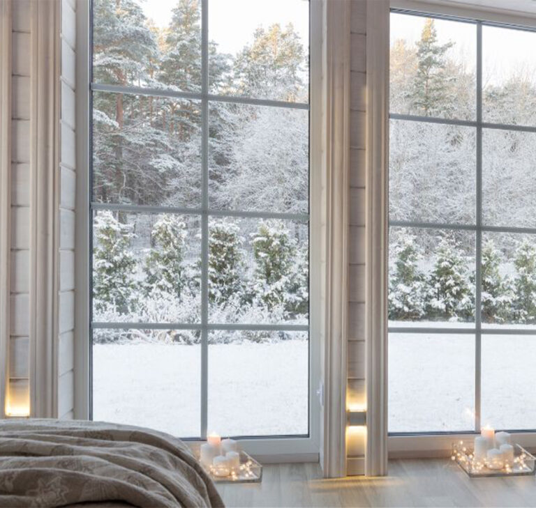 Zoom sur deux baies vitrées en PVC dans une chambre à coucher. À travers les vitres, on distingue un paysage hivernal couvert de neige. À l’intérieur, plusieurs bougies allumées diffusent une lumière douce et orangée, créant une atmosphère calme, intime et cosy malgré le froid extérieur.