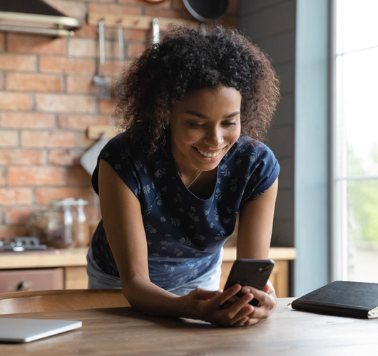 Jeune femme métisse surfant sur son téléphone en souriant dans sa cuisine