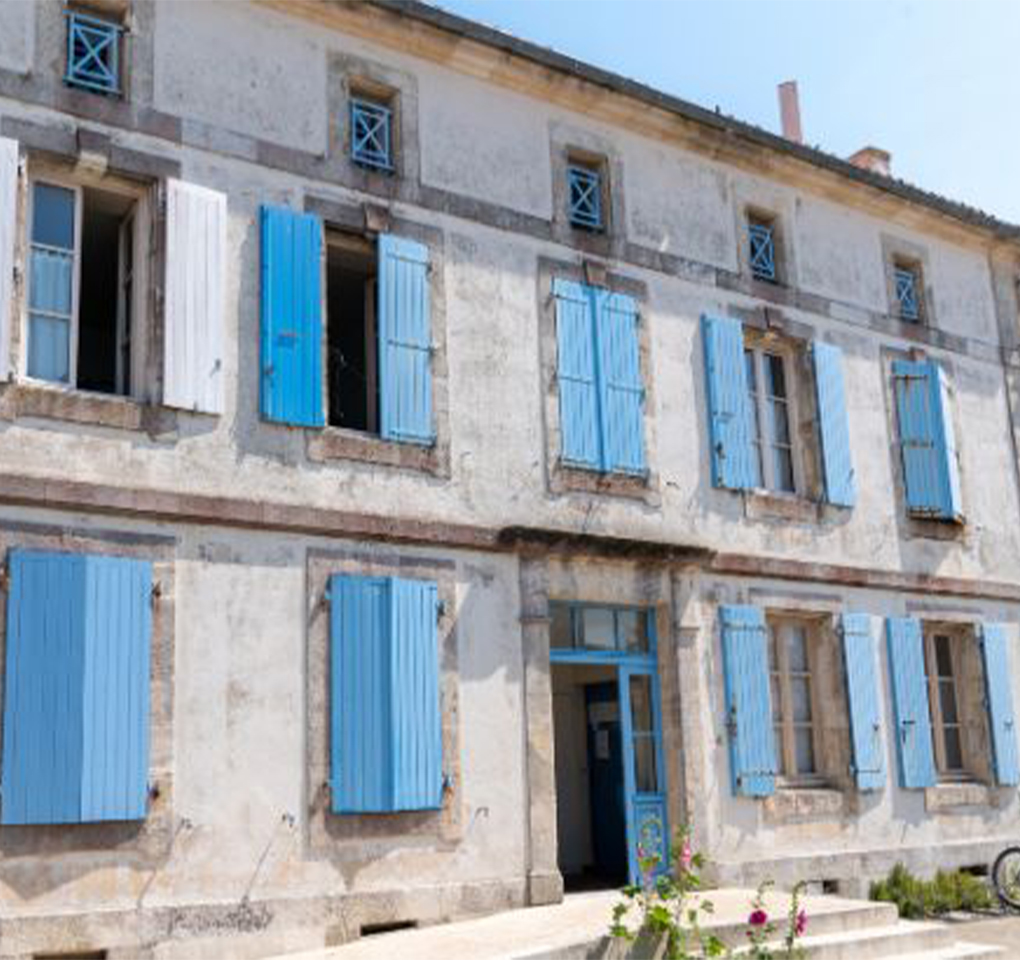 Façade extérieure d’une ancienne grange en béton et pierre, avec volets bleus et anciennes fenêtres à rénover sur toute la façade. 
