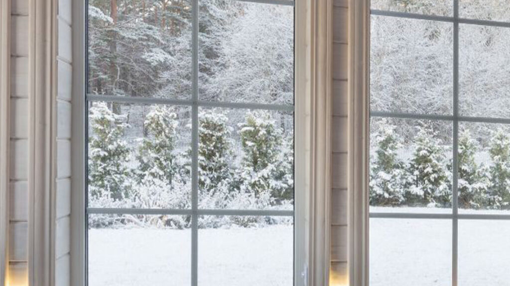 Zoom sur deux baies vitrées en PVC dans une chambre à coucher. À travers les vitres, on distingue un paysage hivernal couvert de neige. À l’intérieur, plusieurs bougies allumées diffusent une lumière douce et orangée, créant une atmosphère calme, intime et cosy malgré le froid extérieur.