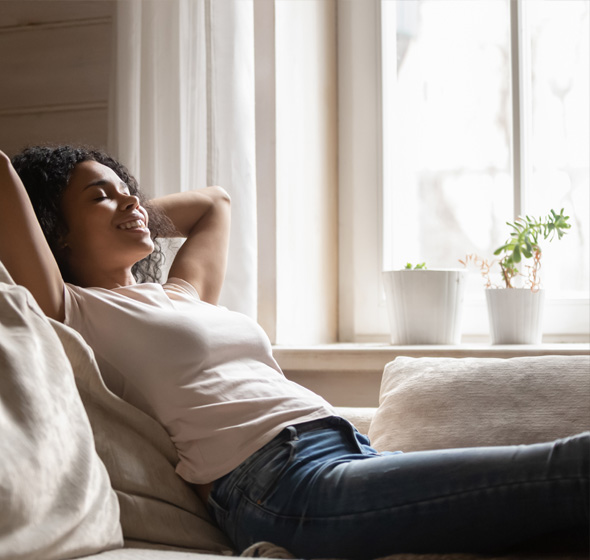 Femme souriante et détendue, installée dans son canapé dans son appartement silencieux grâce à des fenêtres acoustiques amcc